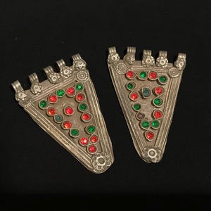 May include: A pair of silver-coloured, triangular pendants adorned with red and green gemstones. Each pendant features a floral design at the base and top, with loops for attachment. The gemstones are arranged in a decorative pattern.