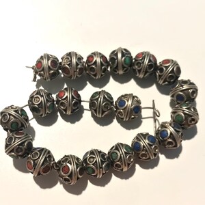 May include: A strand of silver beads with intricate designs and colorful accents. The beads are round and have a textured surface. The colors of the accents are red, green, and blue.