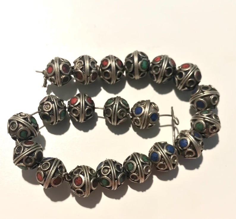 May include: A strand of silver beads with intricate designs and colorful accents. The beads are round and have a textured surface. The colors of the accents are red, green, and blue.
