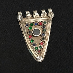 May include: Silver pendant, triangular in shape, featuring a central dark stone and an array of red, green, and blue gemstones. The pendant has loops for suspension and a detailed, ornate design.