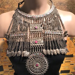 Può includere: Un'elaborata collana argentata con motivi intricati e catene pendenti. La collana presenta ciondoli a forma di foglia, pannelli decorativi con gemme rosa e colorate, indossata sopra un top nero.