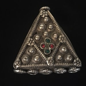 Puede incluir: Un colgante triangular de plata con un diseño decorativo de círculos y flores en relieve. El colgante tiene un diseño central de cuatro piedras preciosas rojas y verdes.