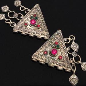 Könnte beinhalten: Zwei silberne dreieckige Anhänger mit roten und grünen Edelsteinen. Jeder Anhänger hat eine Kette mit kleinen silbernen Anhängern.
