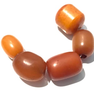 May include: A set of six amber beads in various shades of brown and orange. The beads are oval and round in shape, and they are strung together on a string.