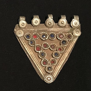Puede incluir: Un colgante triangular plateado con un diseño decorativo. El colgante presenta una serie de piedras preciosas rojas y azules engastadas en un marco de metal detallado. La parte superior tiene cinco pequeños bucles para colgar.