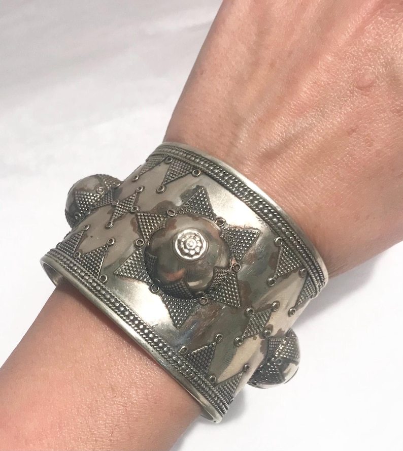 Op de afbeelding: Een zilveren armband met een geometrisch ontwerp en meerdere ronde accenten. De armband is breed en heeft een getextureerd oppervlak.