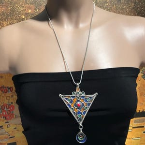 Op de afbeelding: Een zilverkleurige ketting met een driehoekige hanger. De hanger heeft een geometrisch ontwerp met oranje, blauwe, groene en rode accenten. Een druppelvormige bedel hangt eronder. De ketting wordt gedragen op een zwarte strapless top.