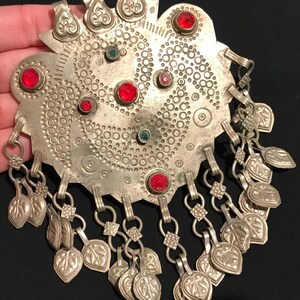 May include: A silver pendant with detailed patterns and red and green gemstone embellishments. The pendant features a central floral design, with hanging leaf-shaped charms. The piece is likely vintage or antique.