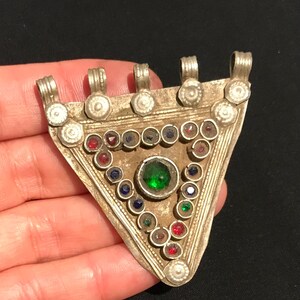 Puede incluir: Un colgante triangular plateado con una piedra verde central y una variedad de gemas de colores. El colgante tiene un diseño detallado con pequeñas decoraciones redondas y bucles para colgar. La pieza se sostiene en una mano sobre un fondo oscuro.