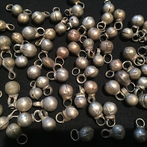 May include: A collection of silver-toned jingle bells with a rounded shape and a small loop at the top. The bells are scattered on a black surface.