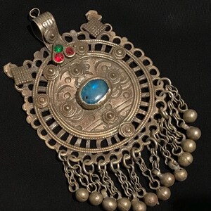 Könnte beinhalten: Verzierter Silberanhänger mit einem zentralen ovalen blauen Stein und kleineren roten und grünen Steinen. Der Anhänger weist aufwendige kreisförmige Designs und baumelnde Ketten mit kleinen Silberperlen auf. Ein dekoratives Stück.