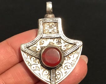 May include: Silver-toned pendant with a shield-like shape, featuring intricate etched designs and a central, round, red gemstone. The pendant has a loop at the top for a chain. The design is ornate and detailed.