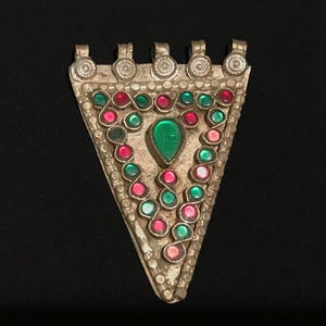 Puede incluir: Colgante triangular plateado con una piedra preciosa verde central y piedras rojas y verdes circundantes. El colgante tiene un diseño detallado con pequeños detalles circulares y lazos en la parte superior para colgar. El fondo es negro.