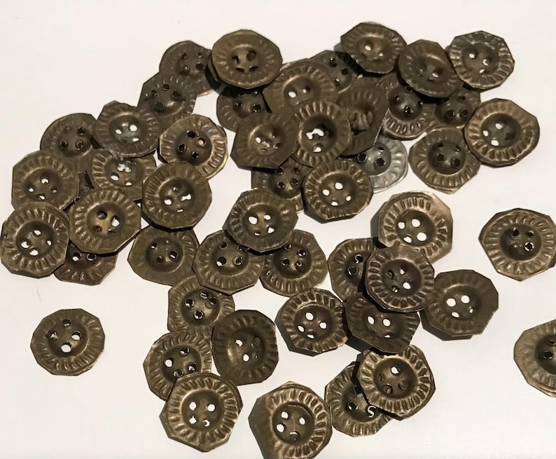 May include: A pile of antique brass-coloured buttons. Each button features a decorative, ridged edge and four holes for sewing. The buttons are scattered on a white surface, highlighting their aged appearance and intricate design.