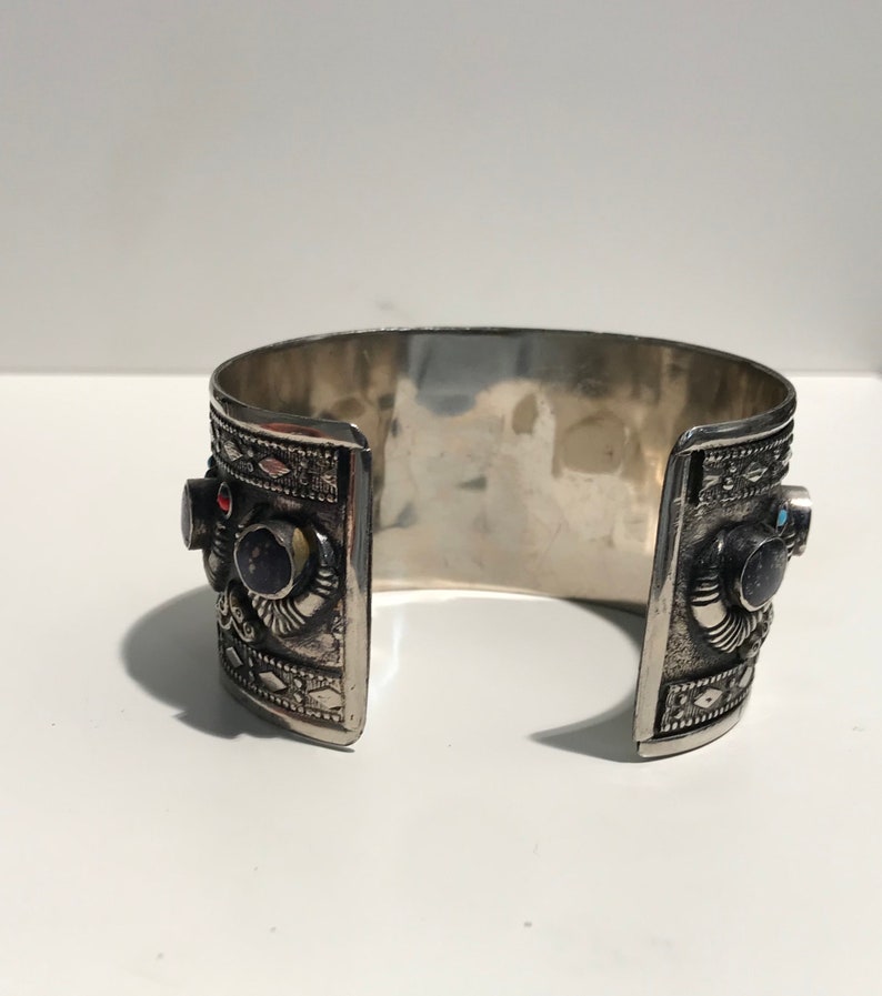 May include: A silver cuff bracelet with intricate designs and colorful gemstones. The bracelet is open on one side and has a hinged closure on the other.