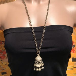 Könnte beinhalten: Silberfarbene Halskette mit langer Kette und glockenförmigem Anhänger. Der Anhänger hat kleine, baumelnde blattartige Anhänger und runde Perlen. Die Halskette wird auf einem schwarzen trägerlosen Oberteil präsentiert.