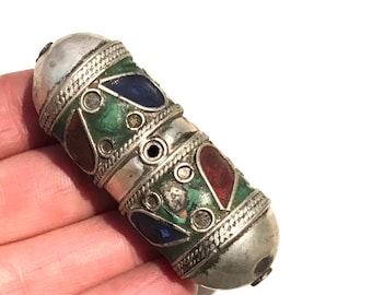 May include: A cylindrical metal bead featuring a silver-toned finish with intricate braided detailing. The surface is inlaid with teardrop-shaped red, blue, and green stone accents, interspersed with small circular metal studs and a central hole.