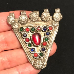 Puede incluir: Colgante triangular plateado con una piedra preciosa roja central y gemas coloridas circundantes. El colgante presenta detalles florales y bucles para colgar. Una pieza de estilo vintage.