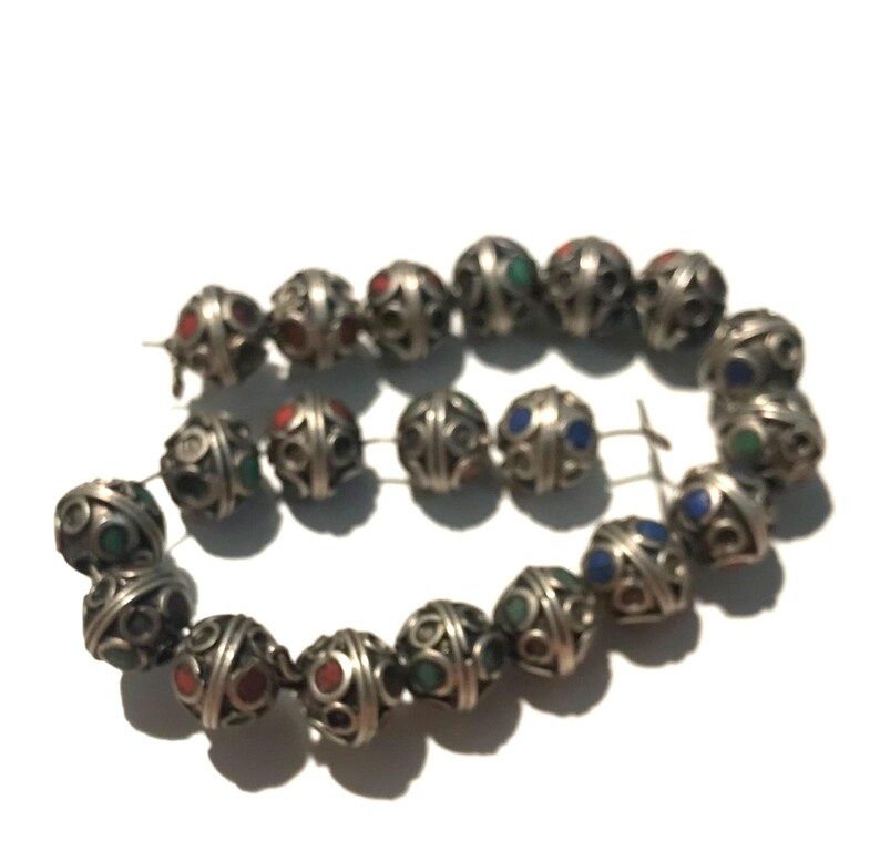May include: A strand of silver beads with intricate designs and colorful inlays. The beads are round and have a slightly irregular shape. The colors of the inlays are blue, green, and red.