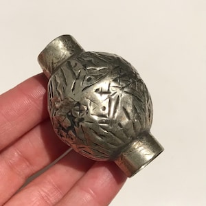 May include: A silver-coloured, decorative bead with an etched, textured surface. The bead has a rounded, bulbous centre and cylindrical ends. The bead is held in a hand against a white background.