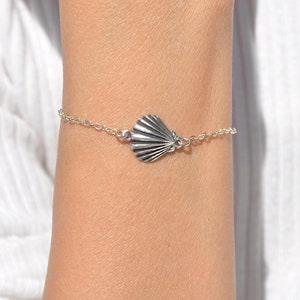 May include: A silver bracelet with a seashell charm. The bracelet is on a person's wrist.