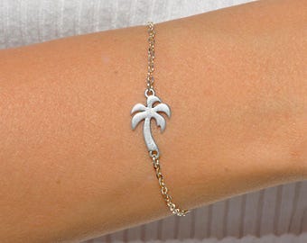 May include: A silver-colored palm tree charm bracelet. The charm is a detailed cutout of a palm tree. The bracelet chain is a delicate gold color. The bracelet is worn on a person's wrist.