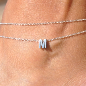 May include: A delicate silver anklet with a small letter "M" pendant. The anklet is composed of two fine chains, creating a layered effect. The jewelry is worn on the ankle, showcasing a minimalist and elegant design.