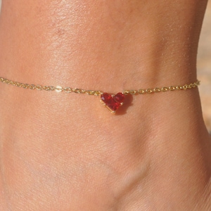 May include: A delicate gold-tone chain anklet featuring a single, small, faceted red heart-shaped charm centered on the chain. The anklet is constructed with fine, interlocking metal links that secure around the ankle with a simple clasp mechanism.