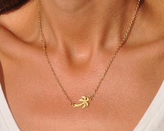 May include: A gold-colored necklace featuring a small palm tree pendant. The necklace is worn around the neck, with the pendant resting near the collarbone. The chain and pendant appear to be made of metal.