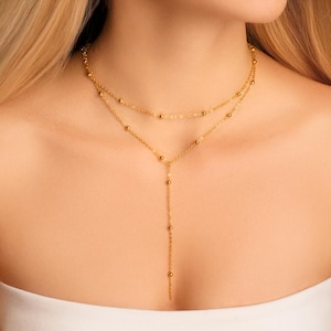 May include: A layered gold necklace with a delicate chain and small, evenly spaced beads. The necklace features a Y-shaped pendant that hangs down the center. The jewelry is displayed on a person's chest.