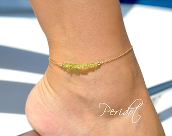 May include: A delicate gold chain anklet with a row of small, light green peridot stones. The anklet is shown on a person's ankle.