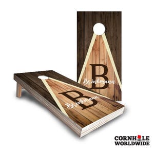 May include: A wooden cornhole board with a brown and beige woodgrain design. The board has a triangle shape with a white circle in the center. The text "Brinkmann" is written in white cursive letters below the triangle. The letter "B" is in a brown circle in the center of the triangle.