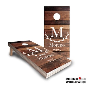 May include: A wooden cornhole board with a rustic woodgrain design. The board is personalized with a white letter "M" in a wreath, the last name "Moreno", and the year "2025".