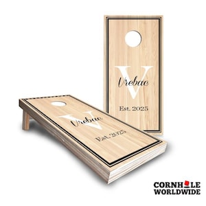 May include: A personalized wooden cornhole board with a black and white border. The board features a white letter "V" and the text "Vrebac Est. 2025".