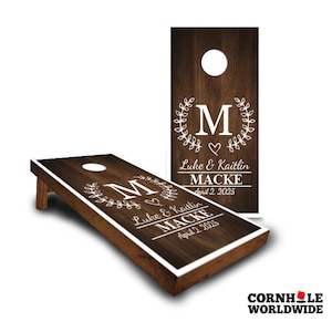 May include: Two dark brown wooden cornhole boards featuring a white circular hole at the top. The surface displays a white printed design with a large letter M, a laurel wreath, a heart, and the text Luke & Kaitlin MACKE April 2 2025.
