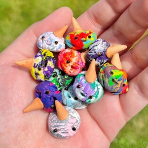 May include: A collection of colorful, miniature ice cream cone sculptures. Each sculpture features a round, textured ice cream scoop with a smiling face and a small, brown cone. The ice cream scoops are various colors, including blue, purple, red, and green.