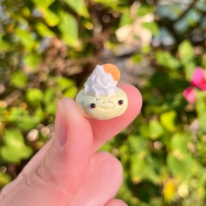May include: A small, whimsical sculpture of a smiling, light yellow creature with black speckles and dark eyes. It has a white, textured topping and a small, orange disc. The sculpture is held between fingers against a blurred green and pink background.