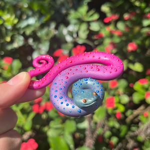 May include: A coiled snake figurine featuring a gradient color scheme that transitions from a vibrant pink with red raised dots to a deep purple and blue with blue raised dots, complete with small orange eyes and a curled tail end.