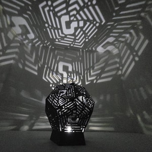 May include: A black metal geometric lamp with a light inside casts a patterned shadow on the wall. The lamp is shaped like a dodecahedron and has a geometric design.