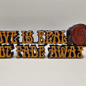 May include: A decorative sign with the words "LOVE IS REAL NOT FADE AWAY" in yellow letters with a dark blue outline. A red rose with green leaves is on the right side of the sign. The sign is on a white surface.