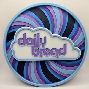 May include: A round, blue and purple swirl design coaster with a white cloud and the text "daily bread" in purple.