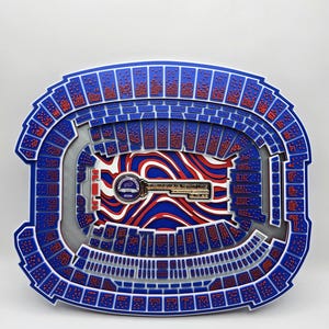 May include: A multi-layered, three-dimensional wall art piece depicting a stadium bowl in blue, white, and red. The center features a metallic key emblem with the text "INAUGURAL SEASON 2023" and a Buffalo Bills logo. The seating sections are detailed with small red dots representing fans.