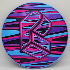 May include: A round, colorful coaster with a 3D geometric design in the center. The coaster has a blue, pink, and purple swirl pattern.