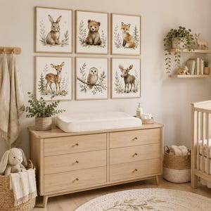 May include: A set of six framed wall art prints featuring watercolor illustrations of a rabbit, bear, fox, deer, owl, and moose, displayed in a nursery with a wooden dresser, a changing pad, a crib, and woven storage baskets.