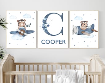 Puede incluir: Tres impresiones artísticas enmarcadas para la guardería. Dos impresiones presentan un oso en un avión azul, nubes y estrellas. La tercera impresión tiene el nombre "Cooper" debajo de una gran letra "C" azul con detalles de estrellas. Las impresiones se exhiben sobre una cuna de madera.