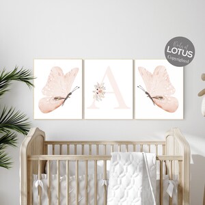 Puede incluir: Tres impresiones de acuarela que representan una mariposa rosa, la letra A con acentos florales rosas y otra mariposa rosa. Las impresiones se muestran en una pared blanca sobre una cuna de madera.