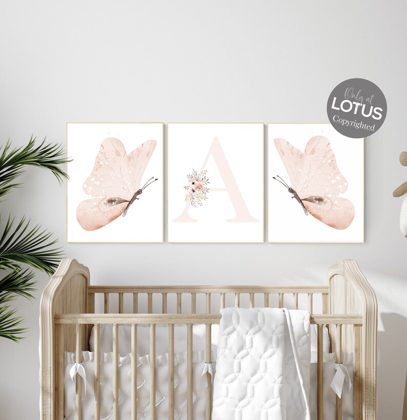 Puede incluir: Tres impresiones de acuarela que representan una mariposa rosa, la letra A con acentos florales rosas y otra mariposa rosa. Las impresiones se muestran en una pared blanca sobre una cuna de madera.
