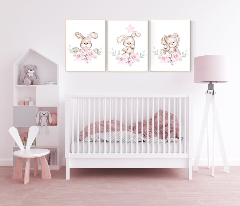 May include: Three framed prints with watercolor illustrations of bunnies with pink flowers. The bunnies are wearing pink bows and have a star on their heads. The prints are hung on a white wall above a white crib.