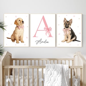 Puede incluir: Conjunto de tres láminas enmarcadas con ilustraciones en acuarela. Una lámina muestra un cachorro de golden retriever con una pajarita rosa. Otra muestra la letra "A" rosa con un lazo y el nombre "Amelia". La tercera lámina representa un cachorro de pastor alemán con un lazo rosa.