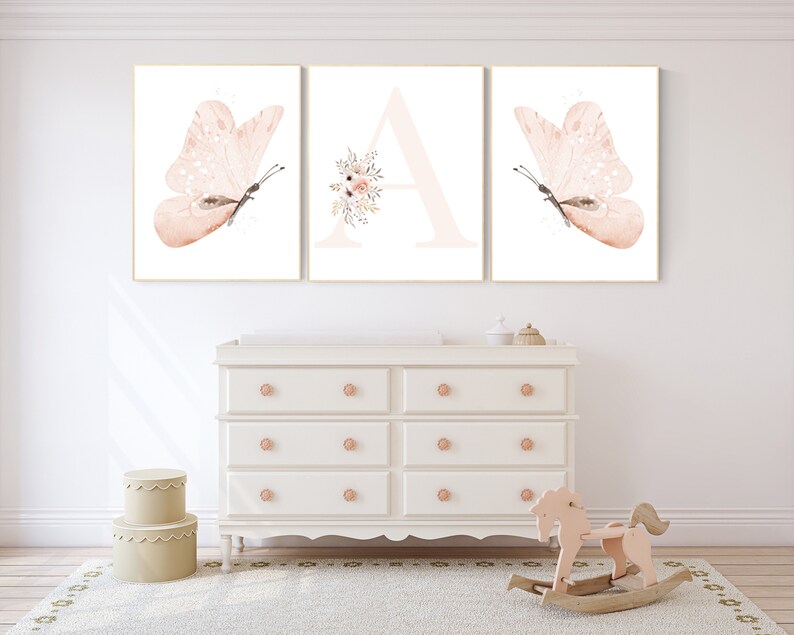 Puede incluir: Tres impresiones enmarcadas con un dise&ntilde;o de acuarela de una mariposa, la letra A con acentos florales y otra mariposa. Las impresiones se muestran en una pared blanca sobre una c&oacute;moda blanca.